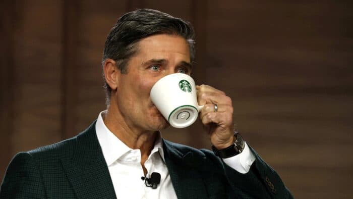 Alt Text: Brian Niccol, Starbucks CEO, drinking from a white Starbucks ceramic mug while wearing a dark green checkered blazer and a lapel microphone.