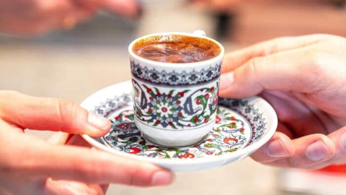 Two hands holding a traditional, ornate Turkish coffee cup and saucer filled with freshly brewed coffee