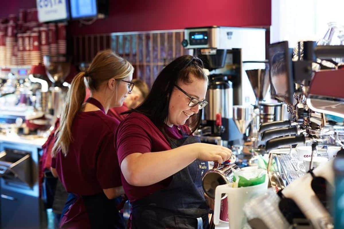 Two Costa Coffee baristas in matching uniforms prepare drinks behind the counter, with one pouring coffee and smiling while working at the espresso machine.