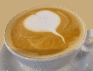 A close-up of a cup of cappuccino with smooth latte art in the shape of a heart, served in a white cup and saucer against a light brown background