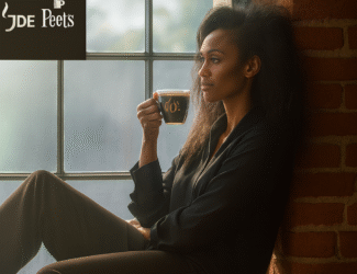 A woman with voluminous curly hair sits on a windowsill in a cozy, brick-walled room, gazing thoughtfully outside while holding a glass cup of coffee. She is dressed in dark, relaxed clothing, and warm natural light filters through the large window. The JDE Peet’s logo appears in the upper left corner, while the beige banner below reads: “Condensed Consolidated Unaudited Interim Financial Statements for the Six-Month Period Ended 30 June 2025,” accompanied by brand logos like Peet’s, Jacobs, and others.