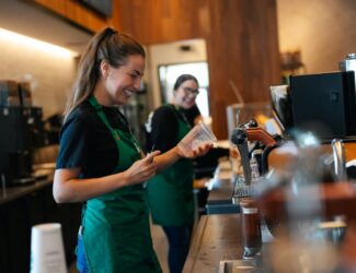 Two Starbucks baristas wearing green aprons are smiling and preparing drinks behind the counter. One holds a clear plastic cup and a pen, while the other smiles in the background. The café interior features warm wood accents and coffee equipment.