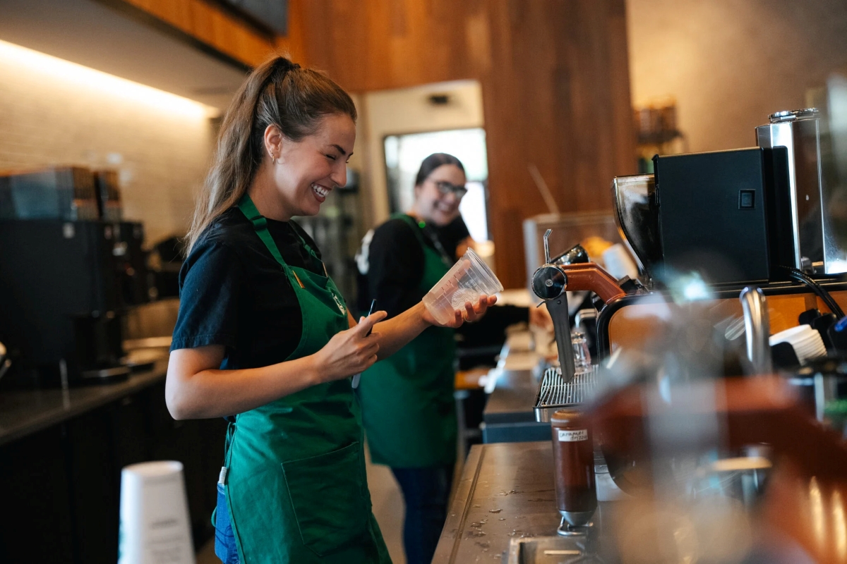 Two Starbucks baristas wearing green aprons are smiling and preparing drinks behind the counter. One holds a clear plastic cup and a pen, while the other smiles in the background. The café interior features warm wood accents and coffee equipment.