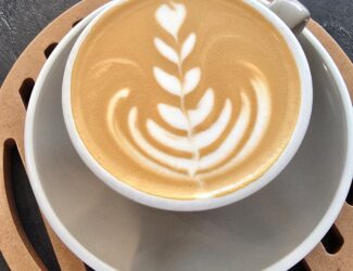 A cup of latte with intricate rosetta-style latte art served on a ceramic saucer over a wooden coaster, placed on a dark stone table.