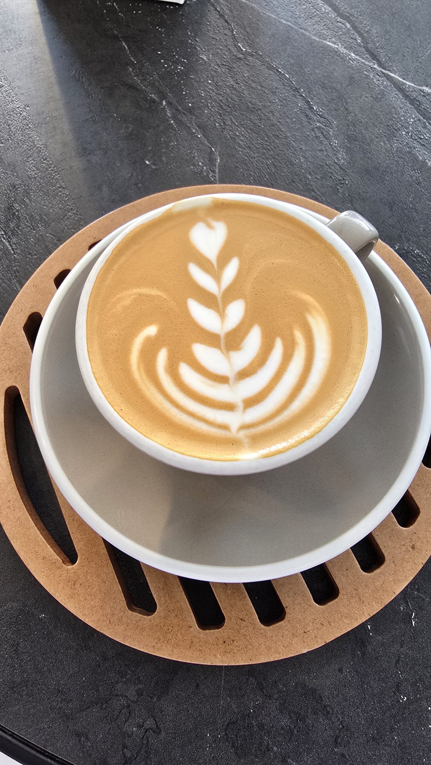 A cup of latte with intricate rosetta-style latte art served on a ceramic saucer over a wooden coaster, placed on a dark stone table.