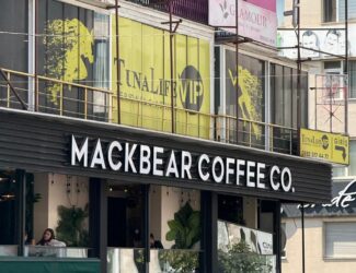 A street view of Mackbear Coffee Co. café in Ankara, located on the first floor above GözGrup Optik and AVON. The café has large windows with customers sitting inside and green plants as interior decor. The building features bright signage and a pink-colored upper level with beauty and fitness businesses, including "TunaLife VIP" and "Glamour Beauty Lounge.