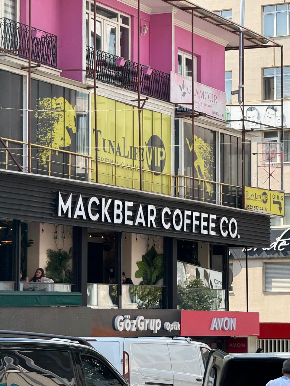 A street view of Mackbear Coffee Co. café in Ankara, located on the first floor above GözGrup Optik and AVON. The café has large windows with customers sitting inside and green plants as interior decor. The building features bright signage and a pink-colored upper level with beauty and fitness businesses, including "TunaLife VIP" and "Glamour Beauty Lounge.