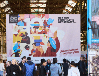 Visitors gather at the Amsterdam Coffee Festival beneath a vibrant banner reading “Hey Hot Stuff! Let’s Get Steamy,” celebrating one decade of specialty coffee culture at NDSM-Loods.