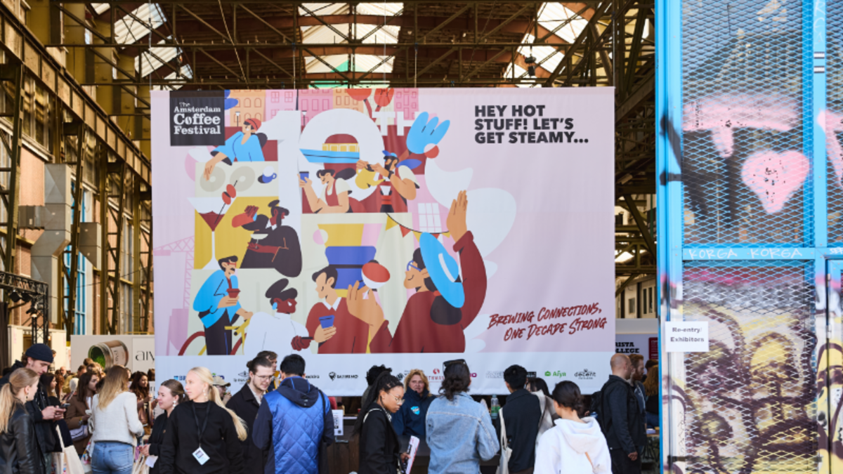 Visitors gather at the Amsterdam Coffee Festival beneath a vibrant banner reading “Hey Hot Stuff! Let’s Get Steamy,” celebrating one decade of specialty coffee culture at NDSM-Loods.