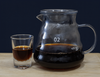 A clear glass carafe and shot glass filled with freshly brewed black coffee on a wooden surface with a clean dark background.
