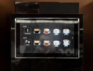 A close-up of a coffee machine touchscreen interface showing drink options and the ability to add nutrients. The left side displays buttons labeled “Add Iodine (I)” and “Add Iron (Fe),” while the right side shows images of various coffee drinks in cups, including espresso, cappuccino, and latte.