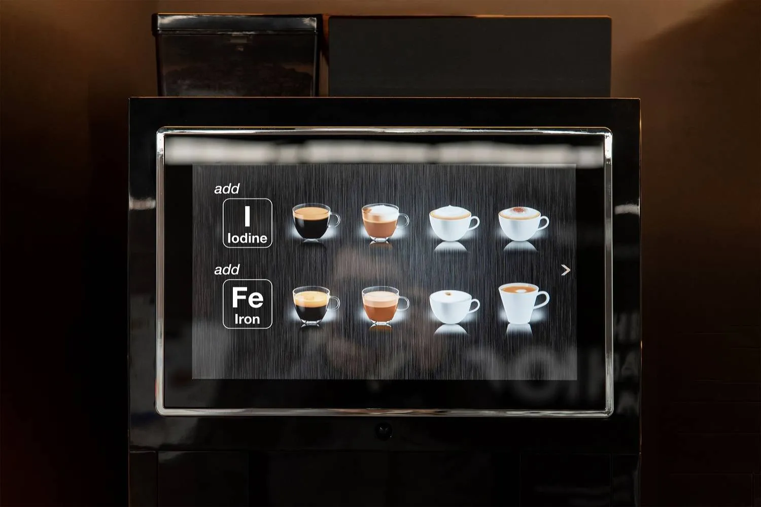 A close-up of a coffee machine touchscreen interface showing drink options and the ability to add nutrients. The left side displays buttons labeled “Add Iodine (I)” and “Add Iron (Fe),” while the right side shows images of various coffee drinks in cups, including espresso, cappuccino, and latte.