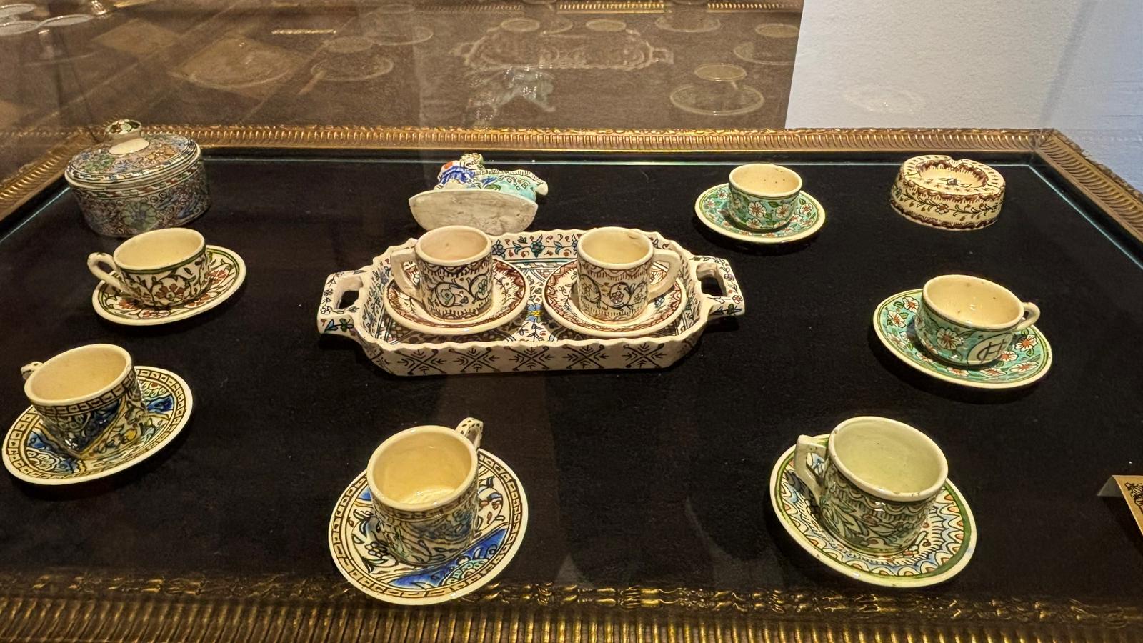 A display case featuring a collection of traditional Turkish coffee cups and saucers, all hand-painted with intricate Ottoman-style floral and geometric patterns. The set includes a decorated serving tray and lidded containers, arranged neatly on a black velvet surface in a museum exhibit.