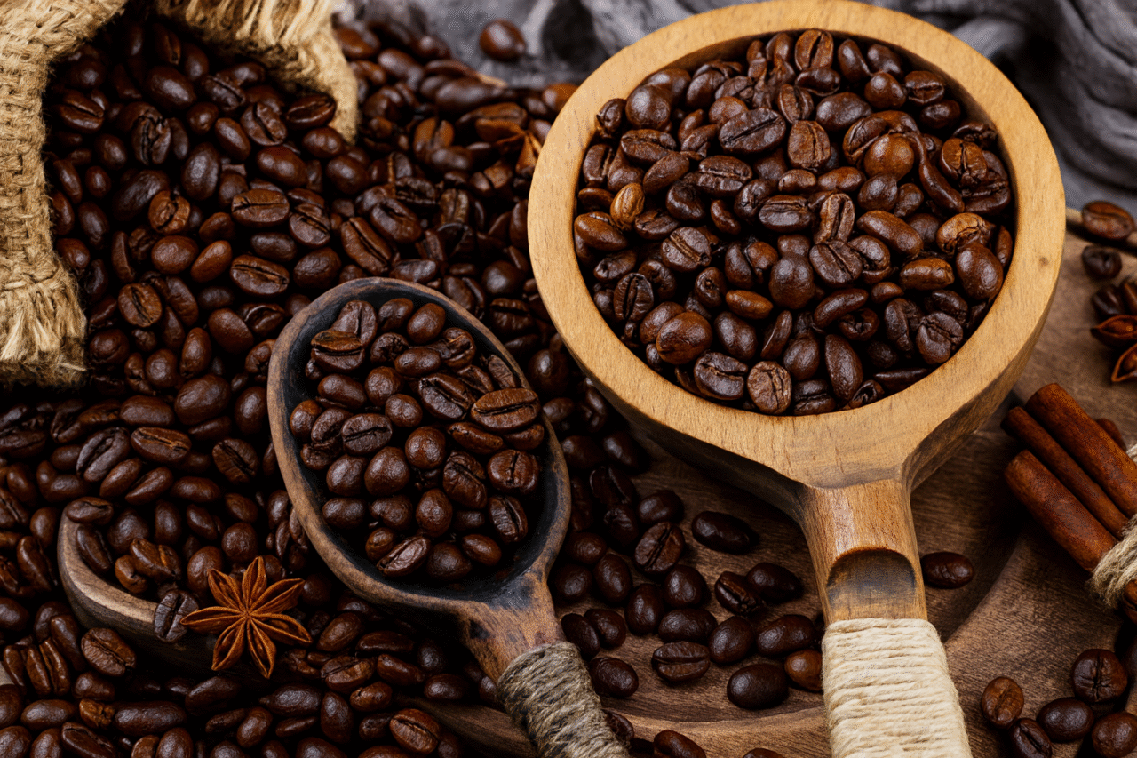A close-up image of dark-roasted Arabic coffee beans spread across a wooden surface, with some beans held in two rustic wooden spoons. A burlap sack spills coffee beans onto the table, and cinnamon sticks and star anise are arranged nearby, enhancing the warm, aromatic atmosphere.