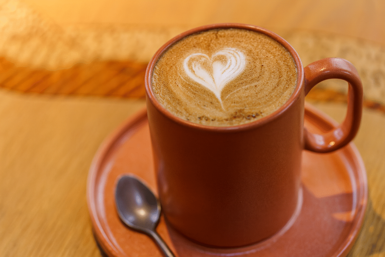 A rust-colored ceramic mug filled with a cappuccino sits on a matching saucer, featuring a heart-shaped latte art design on the frothy surface, with a small spoon resting beside it on a wooden table.