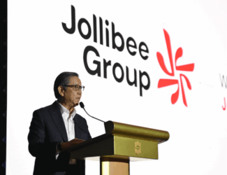 The image shows a man in a dark suit and white shirt speaking at a podium with a microphone. Behind him is a large screen displaying the “Jollibee Group” logo in black text, alongside a red abstract emblem and the tagline “We call this JoyMark.”
