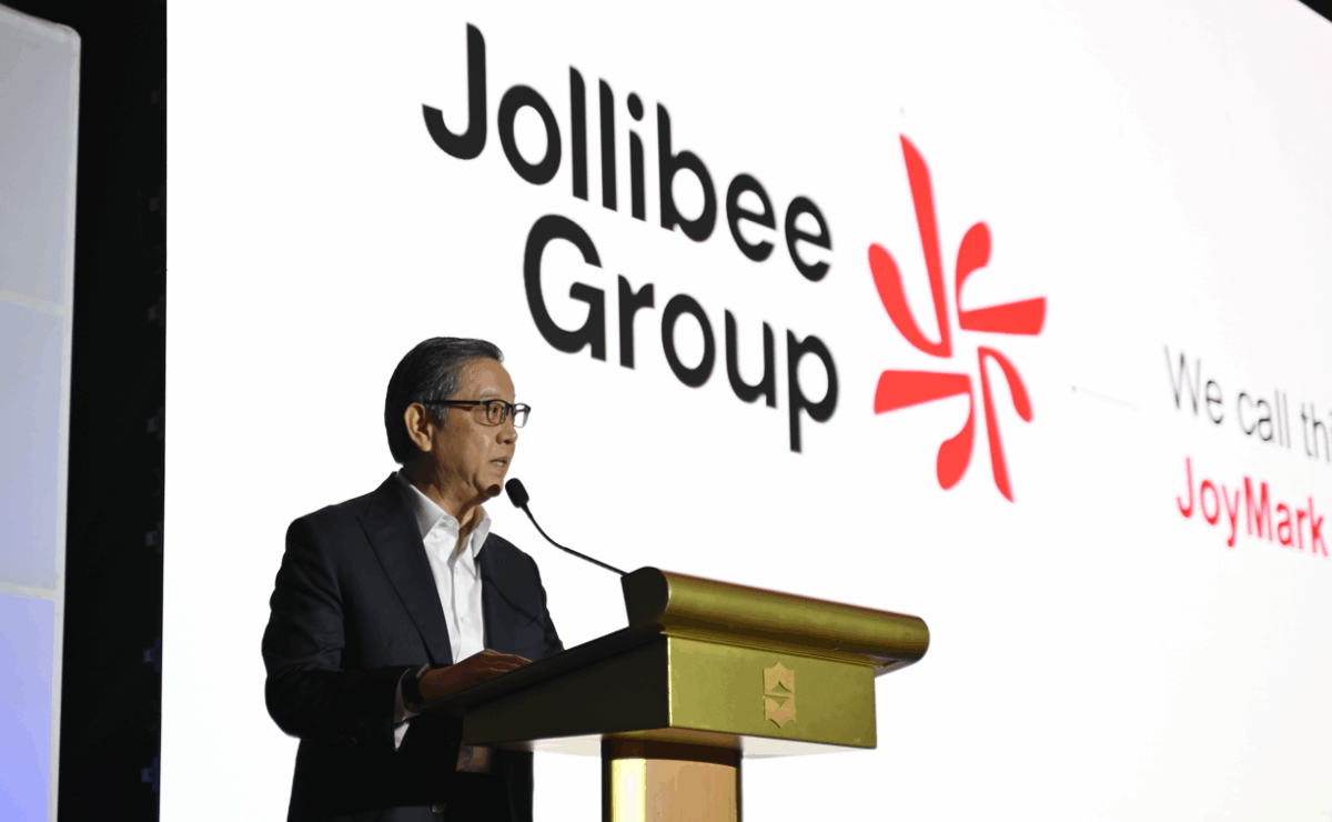 The image shows a man in a dark suit and white shirt speaking at a podium with a microphone. Behind him is a large screen displaying the “Jollibee Group” logo in black text, alongside a red abstract emblem and the tagline “We call this JoyMark.”