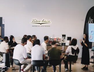 A group of participants attentively listens to a presenter during a coffee science workshop at the Victoria Arduino Experience Lab in Dubai, with coffee machines and a presentation screen in the background.