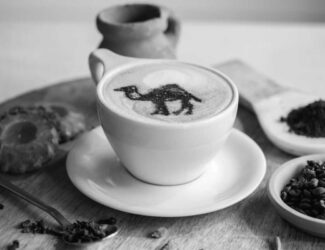 A cup of cappuccino with a camel design in the milk foam, served on a wooden tray alongside coffee beans, ground coffee, a cookie, and a ceramic pot — representing a Middle Eastern or Yemeni coffee setting.