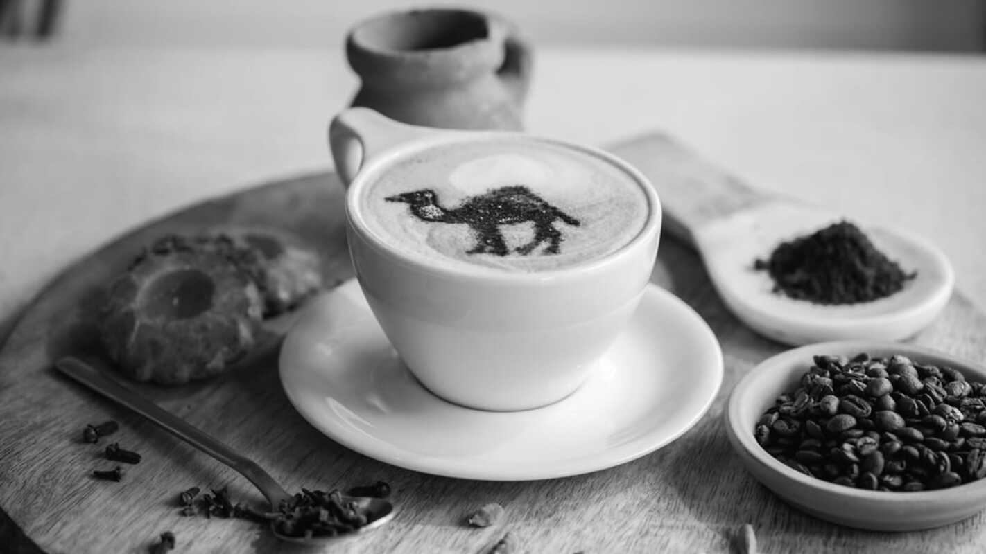 A cup of cappuccino with a camel design in the milk foam, served on a wooden tray alongside coffee beans, ground coffee, a cookie, and a ceramic pot — representing a Middle Eastern or Yemeni coffee setting.