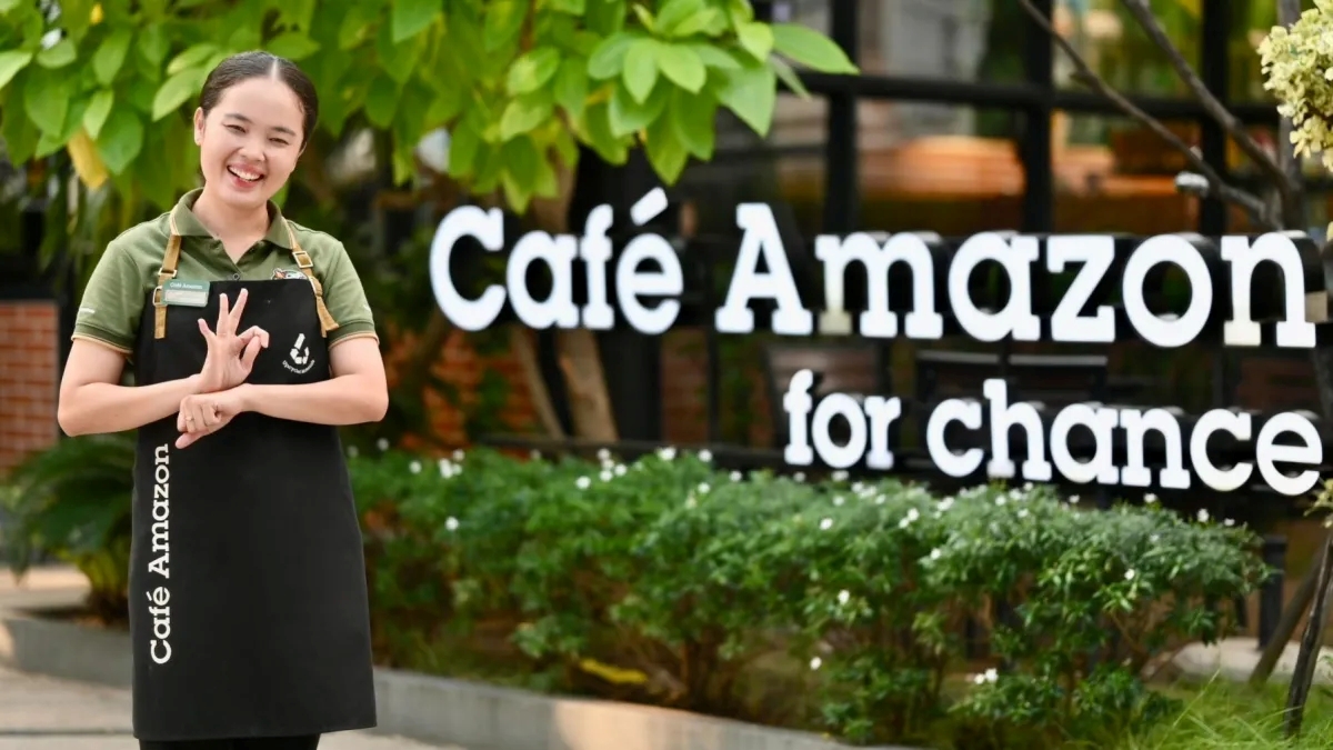 A smiling barista wearing a "Café Amazon" apron stands in front of a sign that reads "Café Amazon for chance." She is using sign language, indicating the café’s inclusive hiring program. Green plants and trees surround the entrance, creating a welcoming atmosphere.