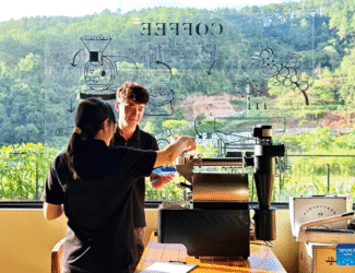 Two staff members roast coffee beans at a café in Nandaohe Village, Pu’er, Yunnan Province, with green hills visible through the window.