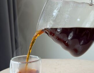A hand pours steaming black coffee from a glass carafe into a clear double-walled glass cup on a light marble table.