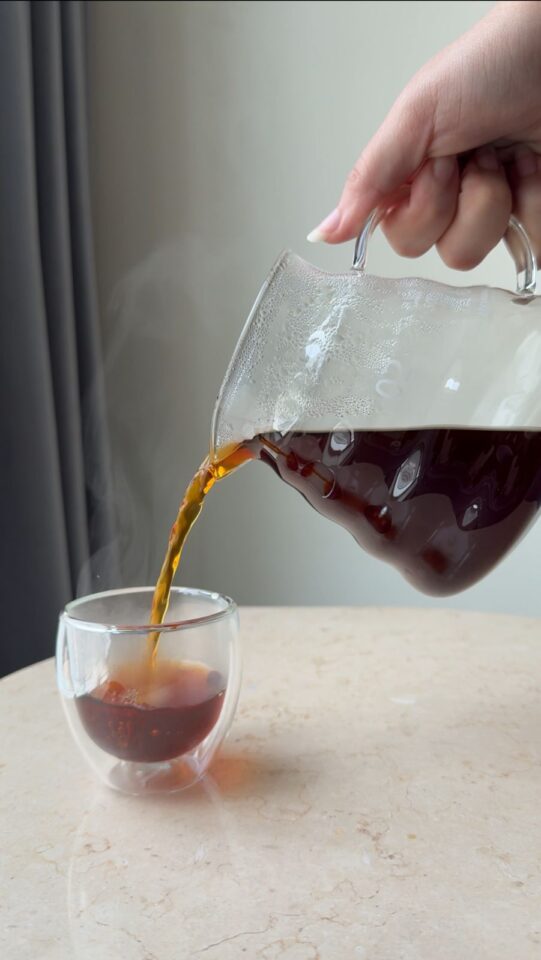 A hand pours steaming black coffee from a glass carafe into a clear double-walled glass cup on a light marble table.