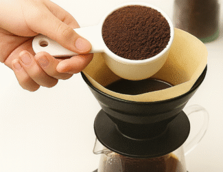 A close-up photo showing a hand holding a white scoop filled with freshly ground coffee, about to pour it into a paper filter placed on a black pour-over dripper set on a digital scale, illustrating precise coffee brewing using the golden ratio.