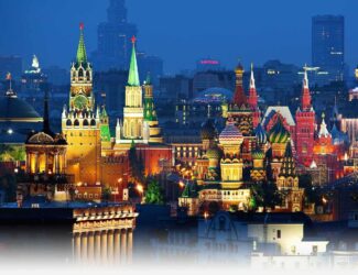 A panoramic night view of central Moscow featuring the brightly illuminated Kremlin towers, Saint Basil’s Cathedral, and historic city buildings under a deep blue sky
