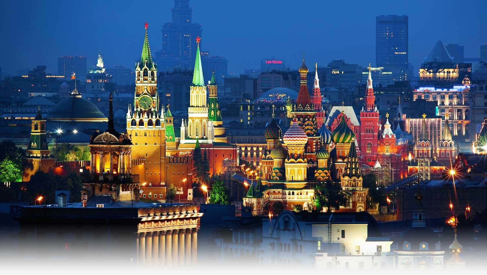 A panoramic night view of central Moscow featuring the brightly illuminated Kremlin towers, Saint Basil’s Cathedral, and historic city buildings under a deep blue sky