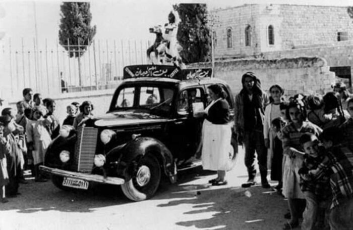 A historical black-and-white photo from Jerusalem showing a vintage car bearing the “Izachman Coffee” sign surrounded by people and children — a glimpse into the early legacy of Palestinian coffee culture.
