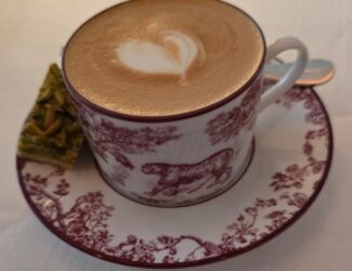 A cup of cappuccino with heart-shaped latte art served in a vintage white-and-maroon cup and saucer featuring animal and floral motifs, placed on a white tablecloth beside a small green pistachio dessert square.