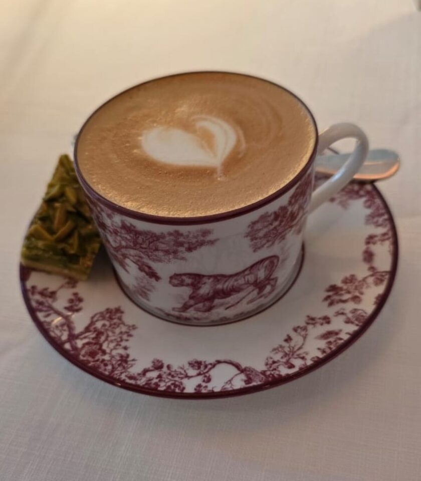 A cup of cappuccino with heart-shaped latte art served in a vintage white-and-maroon cup and saucer featuring animal and floral motifs, placed on a white tablecloth beside a small green pistachio dessert square.