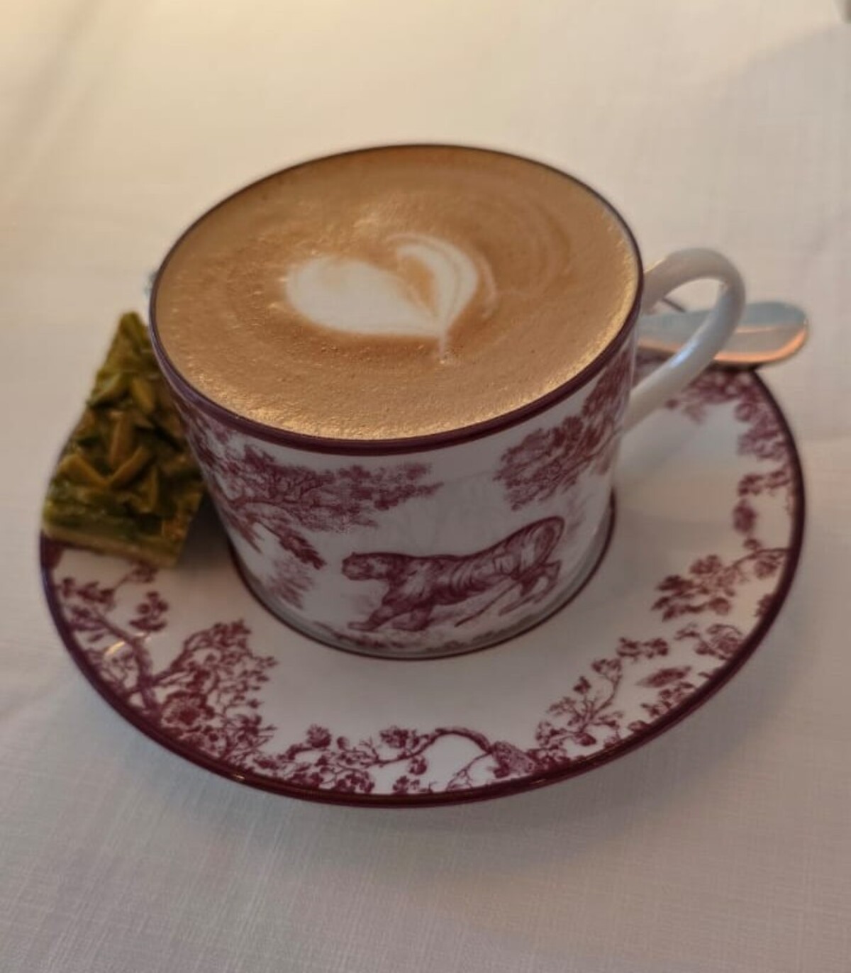 A cup of cappuccino with heart-shaped latte art served in a vintage white-and-maroon cup and saucer featuring animal and floral motifs, placed on a white tablecloth beside a small green pistachio dessert square.