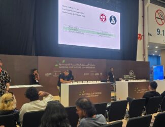 The photo shows the 2025 UAE National Cup Tasters Championship taking place at the Abu Dhabi National Exhibition Centre (ADNEC) during Global Food Week. Judges and participants are seated in front of tasting tables, while a large screen displays competitor names and coffee companies. The event setting includes brown panels with the championship logo and attentive audience members watching the competition.