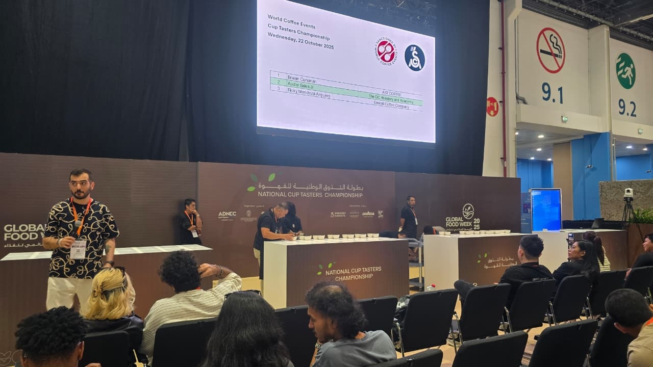The photo shows the 2025 UAE National Cup Tasters Championship taking place at the Abu Dhabi National Exhibition Centre (ADNEC) during Global Food Week. Judges and participants are seated in front of tasting tables, while a large screen displays competitor names and coffee companies. The event setting includes brown panels with the championship logo and attentive audience members watching the competition.