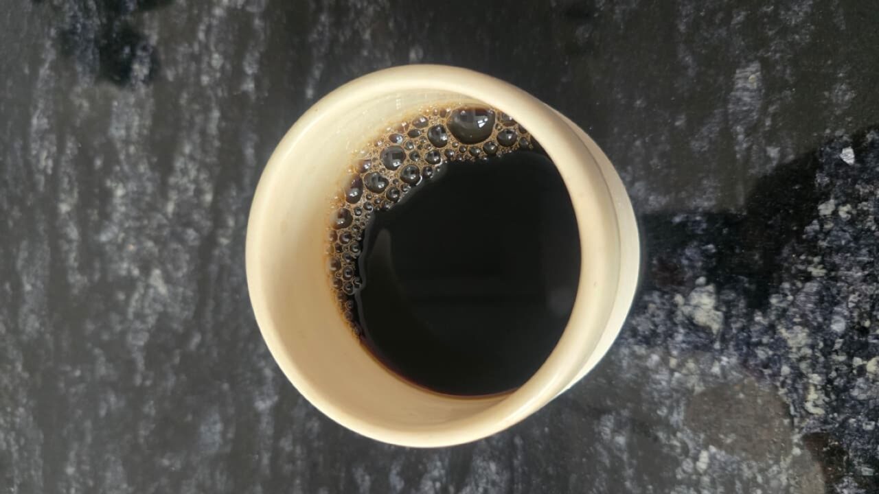 A close-up photo of a cup filled with freshly brewed black coffee placed on a dark granite surface. The crema and small bubbles form along one side of the cup, showing the smooth texture and depth of the coffee