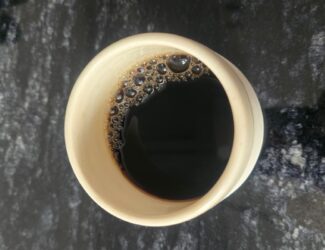 A close-up photo of a cup filled with freshly brewed black coffee placed on a dark granite surface. The crema and small bubbles form along one side of the cup, showing the smooth texture and depth of the coffee