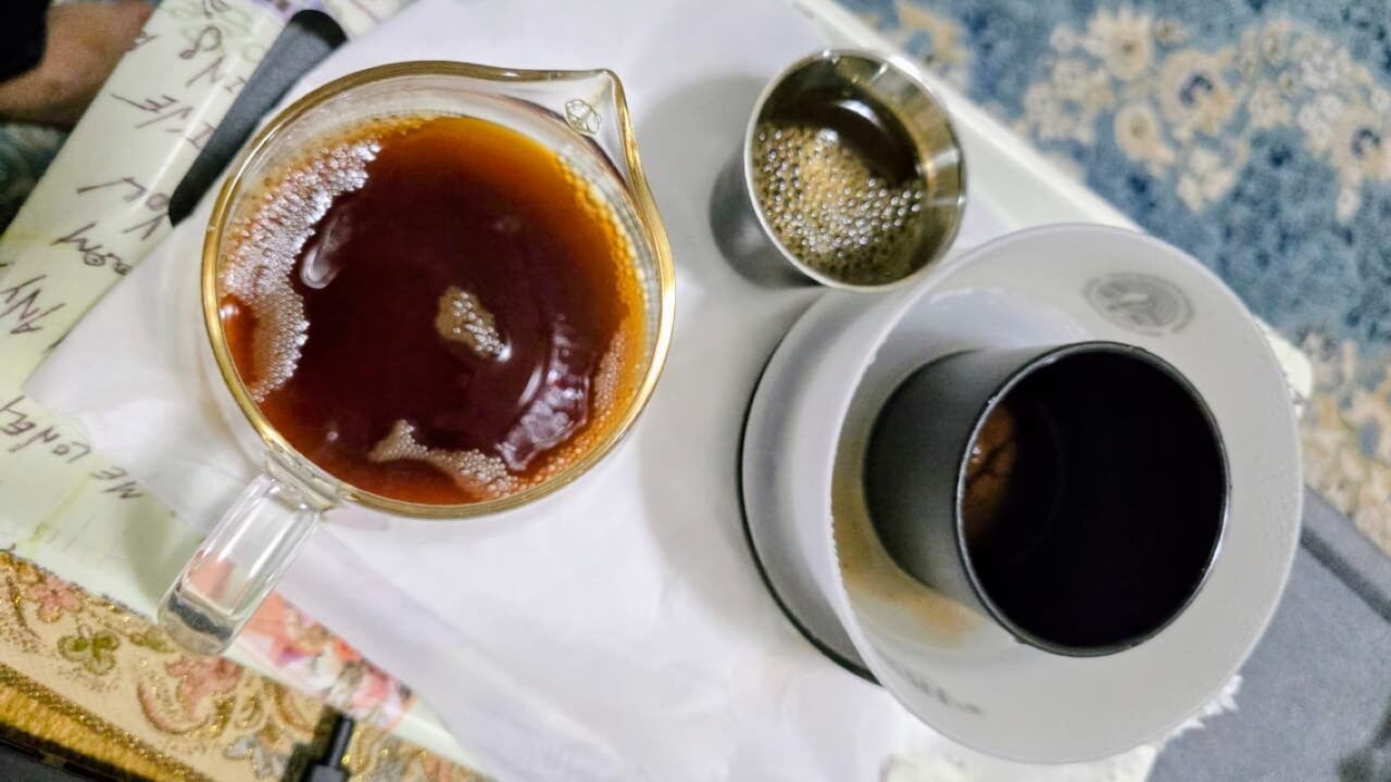 A close-up view of a freshly brewed cup of filtered coffee on a tray. The setup includes a glass serving vessel filled with dark aromatic coffee, a white ceramic dripper with a black inner cone for paper filtration, and a small metal cup with remaining coffee. The image highlights the clean brewing method associated with health-focused coffee preparation.