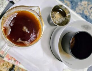 A close-up view of a freshly brewed cup of filtered coffee on a tray. The setup includes a glass serving vessel filled with dark aromatic coffee, a white ceramic dripper with a black inner cone for paper filtration, and a small metal cup with remaining coffee. The image highlights the clean brewing method associated with health-focused coffee preparation.