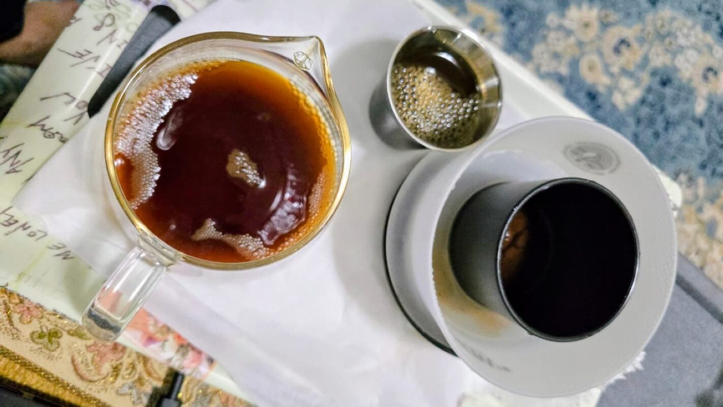 A close-up view of a freshly brewed cup of filtered coffee on a tray. The setup includes a glass serving vessel filled with dark aromatic coffee, a white ceramic dripper with a black inner cone for paper filtration, and a small metal cup with remaining coffee. The image highlights the clean brewing method associated with health-focused coffee preparation.