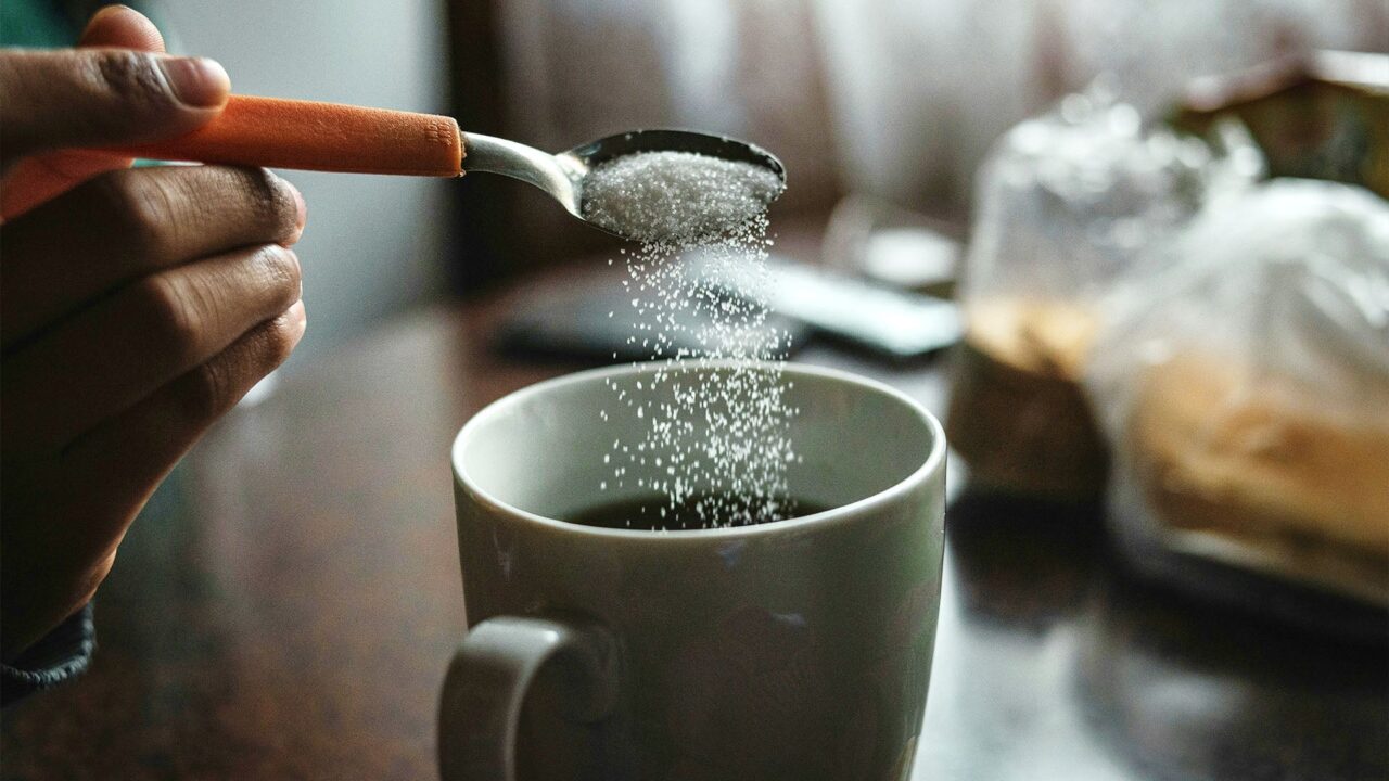 A person holding a spoon pours white granules into a mug of black coffee on a wooden table. The scene is softly lit with natural light, creating a cozy morning atmosphere.