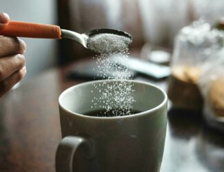 A person holding a spoon pours white granules into a mug of black coffee on a wooden table. The scene is softly lit with natural light, creating a cozy morning atmosphere.