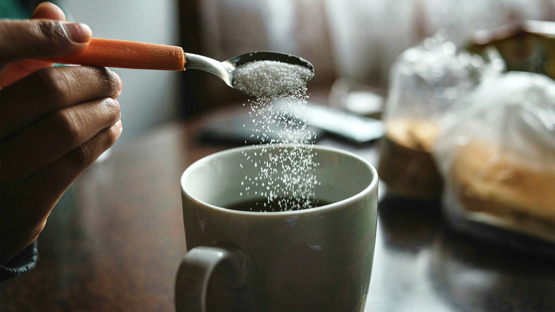 A person holding a spoon pours white granules into a mug of black coffee on a wooden table. The scene is softly lit with natural light, creating a cozy morning atmosphere.