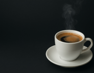 A steaming cup of hot black coffee in a white ceramic cup and saucer placed on a dark background.