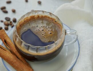 A cup of coffee next to a paper filter and a cinnamon stick on a light background.